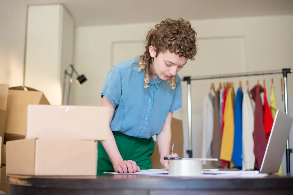 A small business owner working on paperwork beside a laptop, parcels and clothing stock.