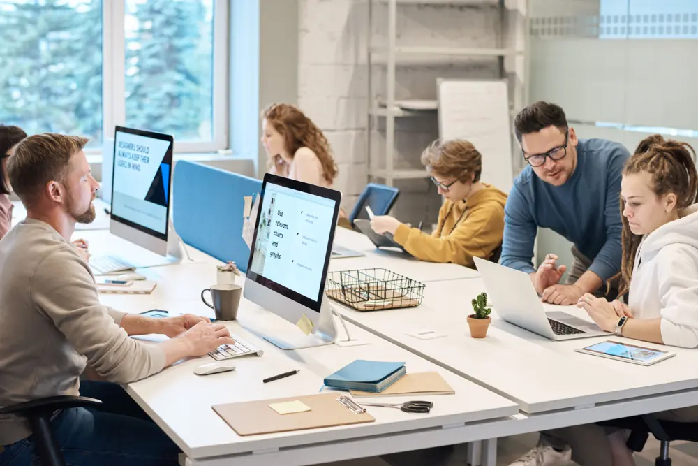 Colleagues working at shared desks with laptops and desktop monitors.
