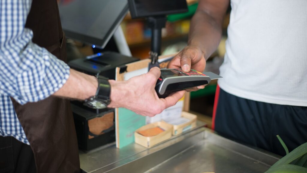 A shopkeeper handing a card payment terminal across the counter to a customer