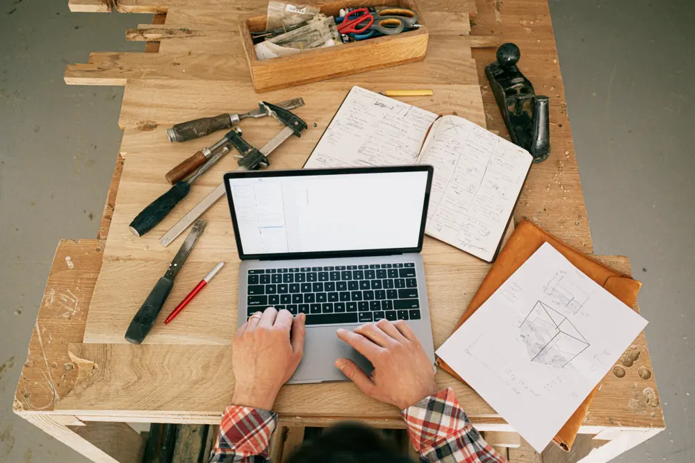 Hands using a laptop on a woodworking bench beside tools and plans.