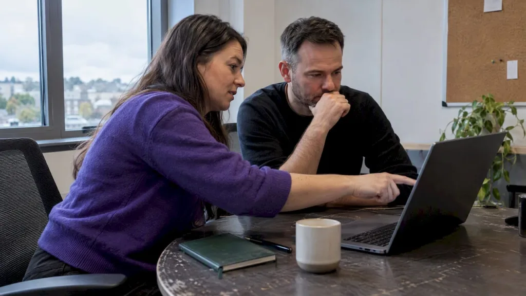 Two colleagues reviewing an open laptop beside a notebook and mugs on an office desk.