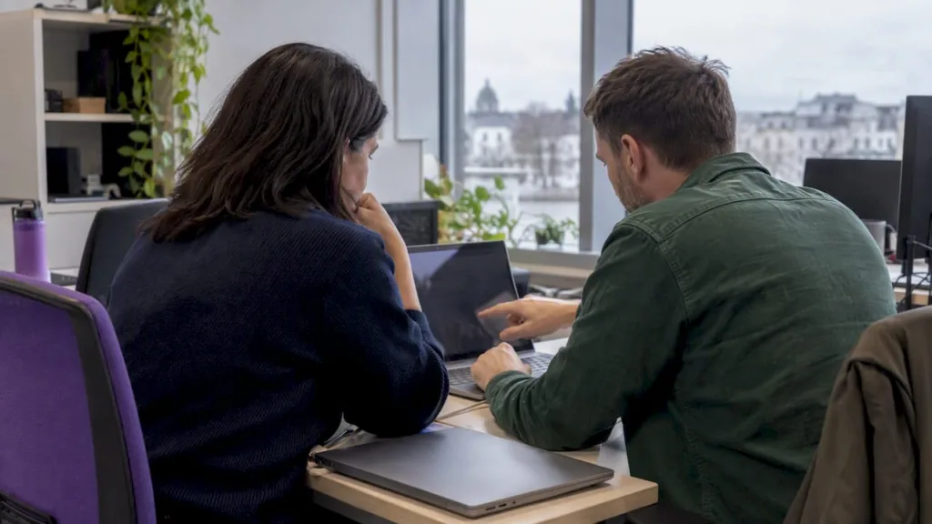 Two colleagues discussing work at a desk with a laptop and desktop monitor.