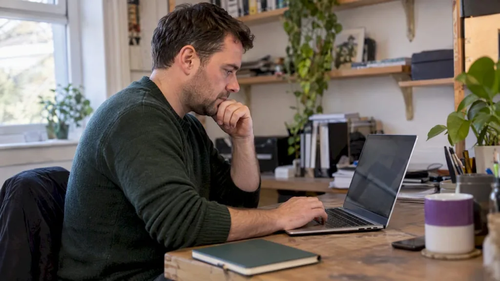 A man using a laptop at a wooden desk with plants and a mug; illustrative image.